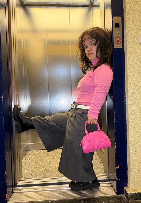 Pantalon gris à jambes larges, haut rose à manches longues, bottes plateformes noires, et un petit sac à main rose dans un ascenseur métallique.