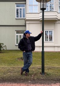 Homme portant une casquette bleue et une veste noire appuyé contre un lampadaire dans une zone herbeuse devant un bâtiment de couleur pâle avec des fenêtres à cadres blancs.