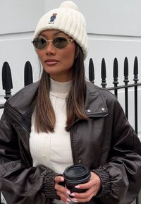 Casaco de couro castanho, suéter branco canelado, gorro de tricot creme com pom-pom e óculos de sol ovais. A segurar uma chávena de café preto.