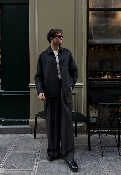 Chaqueta de lana a rayas negras, camisa blanca con cuello debajo, pantalones grises holgados, zapatos negros, de pie frente a un café con grandes ventanales de cristal.