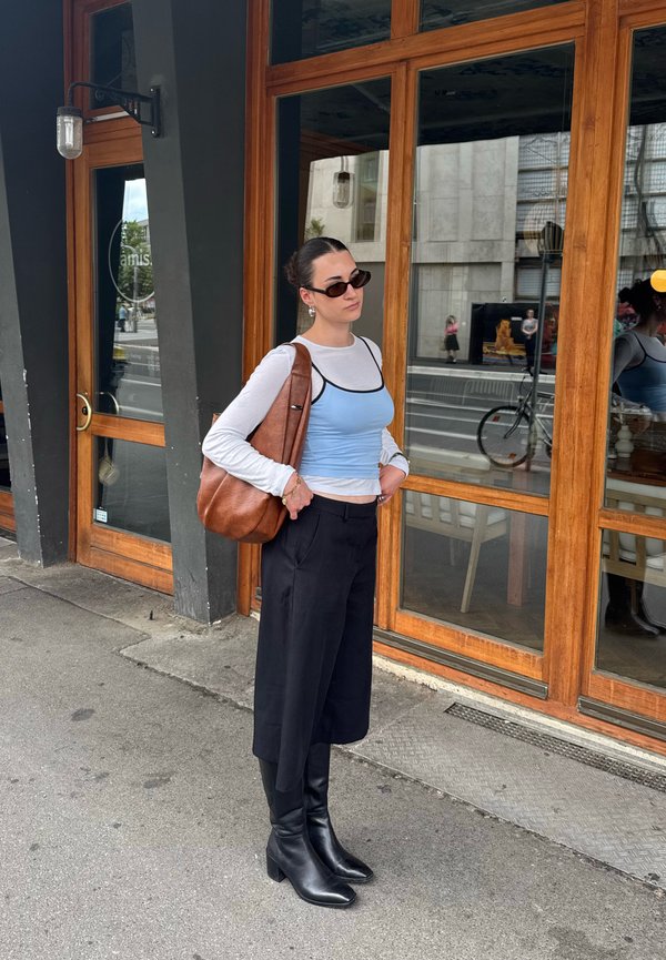 Donna con occhiali da sole, camicia bianca a maniche lunghe sotto una canotta blu, culotte nere e stivali neri, in piedi vicino a una porta di legno con una borsa marrone in mano.