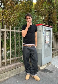 A young adult wearing a fitted black t-shirt, dark baggy jeans, and tan boots stands beside a metal utility box in an urban environment with greenery.