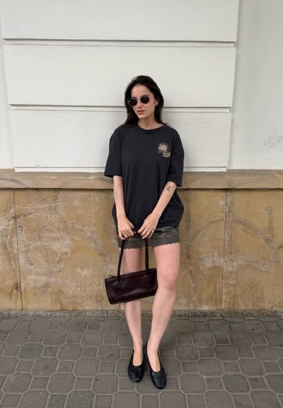 Mujer con camiseta negra de gran tamaño, shorts con encaje, bailarinas negras y gafas de sol sosteniendo un bolso oscuro contra una pared beige.