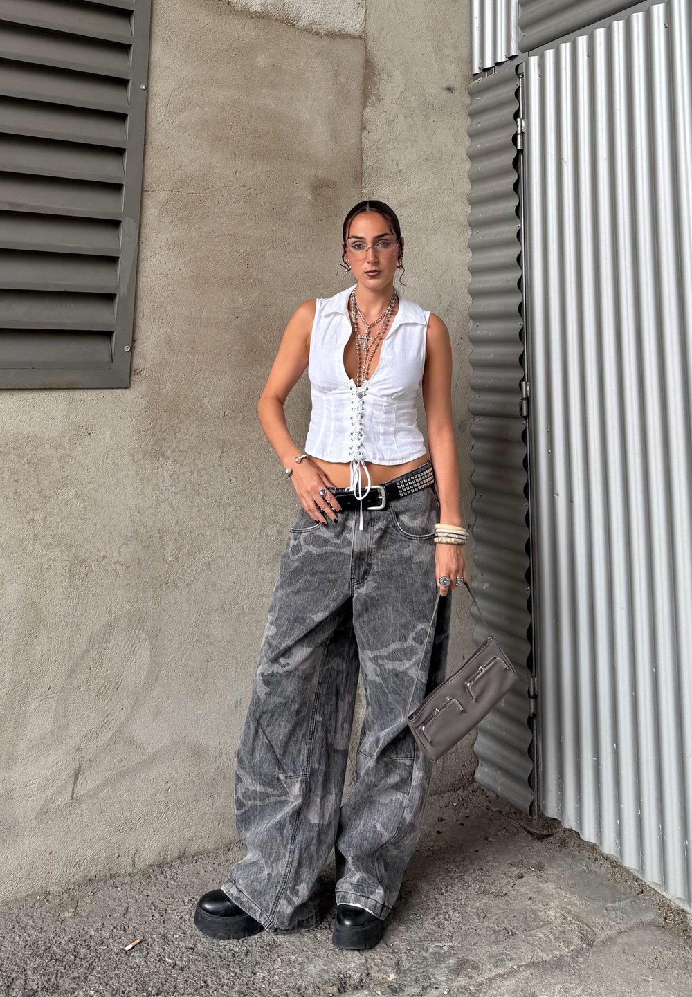 Bílé zkrácené tričko se šněrováním vpředu, šedé oversized kalhoty s vzorem, černé platformové boty a šedá kabelka. Součástí jsou také šperky z různých kovů.