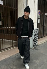 Jeune homme portant un bonnet noir, une veste noire, un jean sombre ample et des baskets blanches, marchant sur un trottoir près d'une entrée grillagée avec des graffitis.