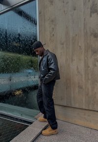 Černá kožená bomber bunda, černé džíny, hnědé pracovní boty, stojící na kamenných schodech před texturovanou skleněnou stěnou s kapkami vody.