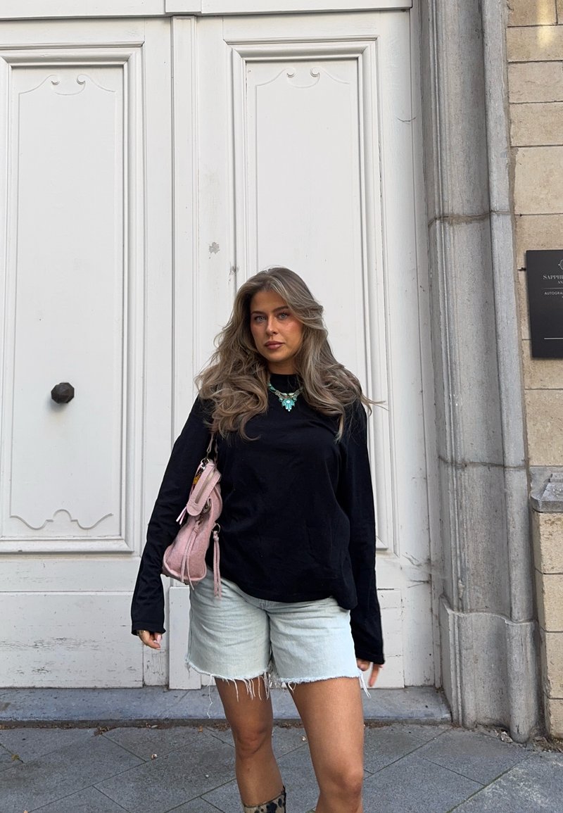 Mujer joven con cabello largo y ondulado lleva puesta una camiseta negra de manga larga, shorts de mezclilla claros, collar turquesa y lleva un bolso rosa, de pie junto a una gran puerta blanca.