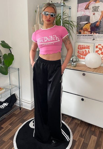 Camiseta rosa corta con logotipo blanco, combinada con pantalones de pierna ancha negros con rayas laterales. El modelo lleva gafas de sol y está de pie en interiores.