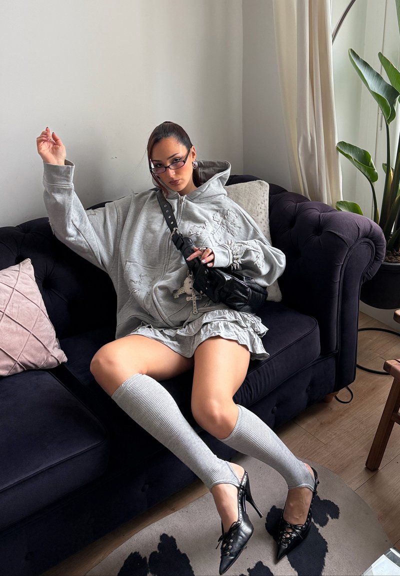 Woman wearing oversized gray hoodie, ruffled skirt, knee-high socks, and black heels sitting on dark couch in living room with plants and cushions.