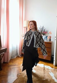 Γυναίκα με κόκκινα μαλλιά, ντυμένη με καρό ζακέτα και μαύρο φόρεμα, περπατά μέσα σε ένα φωτεινό δω μάτιο με ξύλινη τουαλέτα και ροζ κουρτίνες.