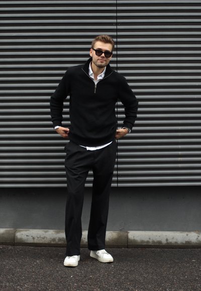 Sudadera de cremallera negra, camisa blanca con cuello debajo, pantalones negros y zapatillas blancas. De pie contra un fondo gris texturizado.