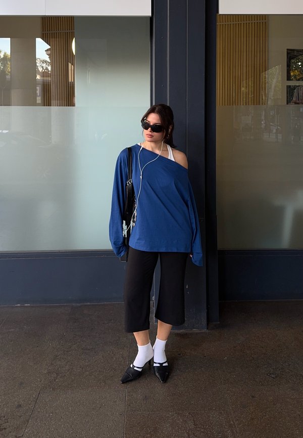 Une femme portant des lunettes de soleil se tient devant un bâtiment à façade vitrée. Elle porte un haut bleu, un pantalon noir court, des chaussettes blanches et des chaussures plates noires.