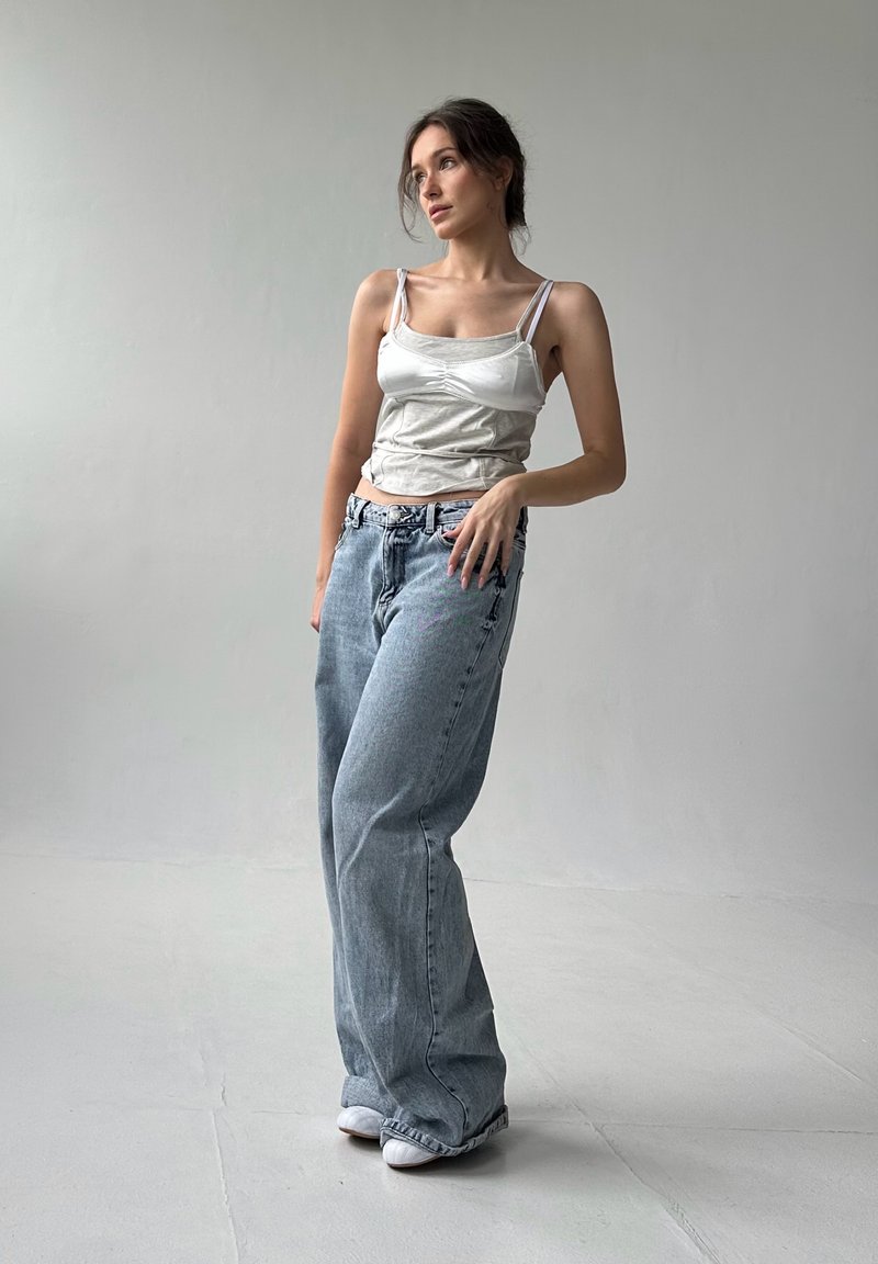 Débardeur blanc à fines bretelles, associé à un jean en denim large et bleu clair et des chaussures blanches, sur fond neutre.