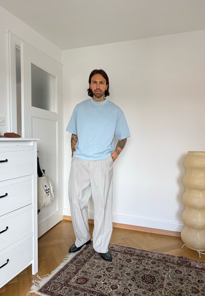 Hombre con tatuajes que lleva una camiseta azul clara y pantalones anchos beige, de pie en una habitación minimalista con paredes blancas y suelo de madera.