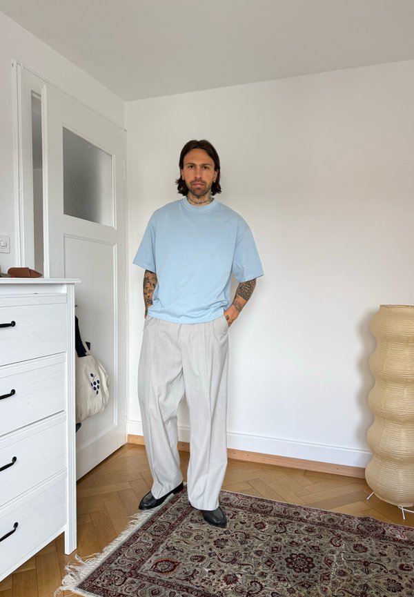 Mann med tatoveringer iført lyseblå t-skjorte og beige vide bukser står i et minimalistisk rom med hvite vegger og tregulv.
