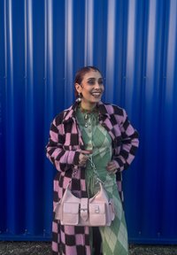 Personne souriante debout contre un mur bleu, portant un manteau à carreaux rose et noir, une tenue verte à motif losange, tenant un sac à main rose clair avec une bandoulière chaîne.