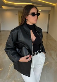 Sort jakke i kunstlæder med krave og lynlås foran, kombineret med en sort rullekrave og hvide bukser med h�øj talje. Holder en lille sort taske.