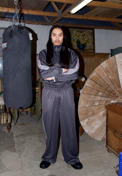 Persona con cabello largo y negro, vestida con chándal gris, de pie con los brazos cruzados junto a un saco de boxeo colgado y una gran rueda de madera en un sótano.