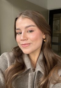 Jonge vrouw met lang bruin haar en gouden oorbellen, gekleed in een beige jas, die binnenshuis voorzichtig glimlacht met een neutrale achtergrond.