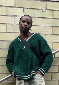 Πράσινο πλεκτό πουλόβερ με V λαιμόκοψη και λευκές ρίγες στους καρπούς. Διαθέτει υφασμάτινο σχέδιο καλωδίων και ένα μικρό κολιέ με μενταγιόν.