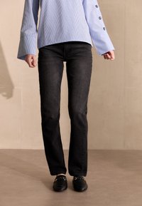 Hellblau gestreiftes Button-Up-Hemd mit Glockenärmel, kombiniert mit schwarzer gerader Jeans und schwarzen Loafers mit einem Goldkettendetail.