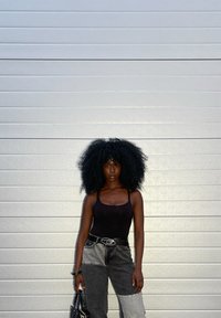 Top nero a canottiera abbinato a jeans grigi a vita alta con tonalità divise. Borsa nera tenuta in una mano. Capelli voluminosi e ricci. Sfondo neutro.