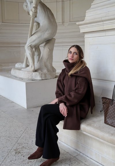 Mujer con abrigo marrón y pantalones negros sentada en un banco de piedra junto a una gran escultura de mármol dentro de la sala de un museo.