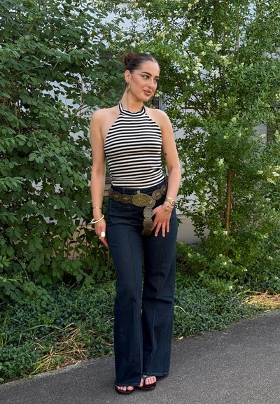 Mujer con un top tipo halter de rayas en blanco y negro, vaqueros acampanados oscuros y cinturón decorativo, de pie en un camino con arbustos verdes detrás de ella.