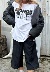 Juodas pūkuotas džemperis virš baltos marškinėlių su užrašu "BLONDIE" ir grafiniu veidų vaizdu. Laidi juoda treniruočių kelnės su baltomis šoninėmis juostelėmis.