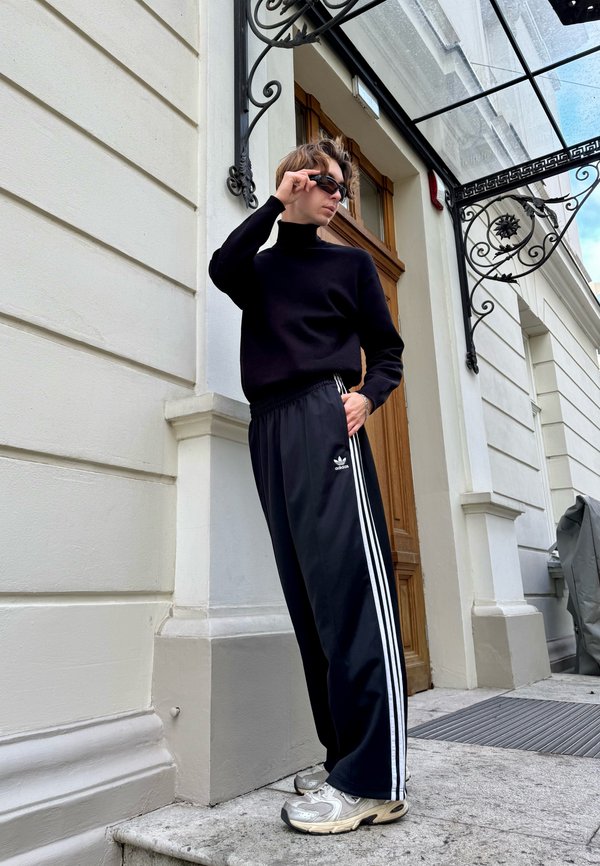 Pulôver de gola alta preto emparelhado com calças de treino pretas da Adidas com listras brancas. Usado com sapatilhas cinzas, apoiado contra um prédio.