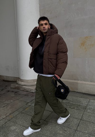 Chaqueta puffer marrón, sudadera negra con capucha, pantalones cargo oliva y zapatillas blancas. Sosteniendo una pequeña bolsa negra de hombro. Ambiente urbano con fondo de piedra.