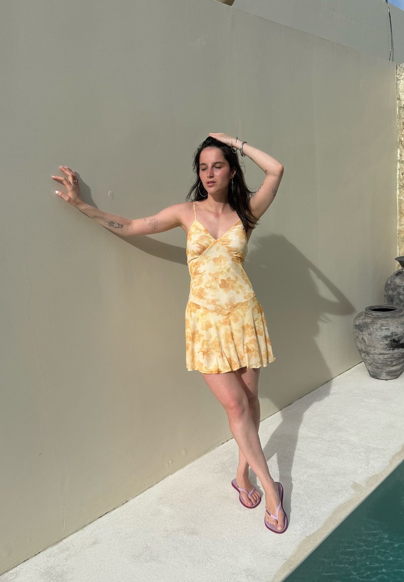 Kvinde i en gul blomstret kjole og flip-flops står ved en beige væg ved siden af en pool, øjnene lukkede, med den ene hånd på hovedet og den anden rørende ved væggen.