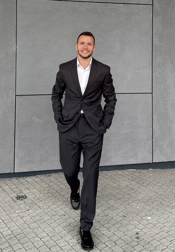 Abito nero con camicia bianca, design su misura, texture liscia e scarpe nere. In piedi su un pavimento grigio testurizzato con sfondo di muro grigio.