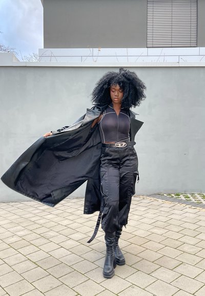 Abrigo de cuero negro tipo trench sobre una blusa negra ajustada con costuras blancas, pantalones cargo y botas negras hasta la rodilla; pelo negro ondulado.