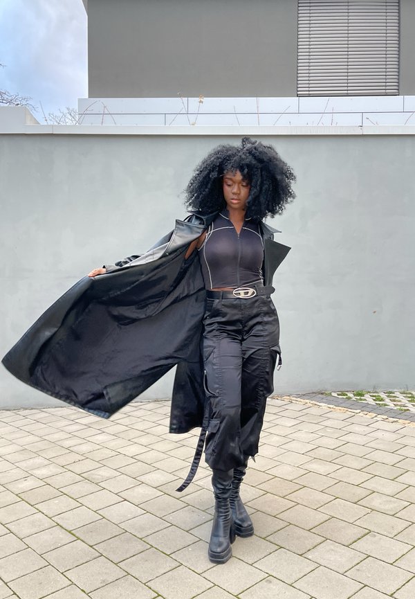 Casaco trench de pele preto sobre uma blusa preta ajustada com costura branca, calças cargo e botas altas pretas; cabelo preto ondulado.