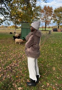 Ruda, pūkuota kailio striukė su pūkuotu apdailu, pilka megzta kepurė, šviesiai pilkos kelnės ir juodi gumbačiai. Fone matyti avys ir žaluma.