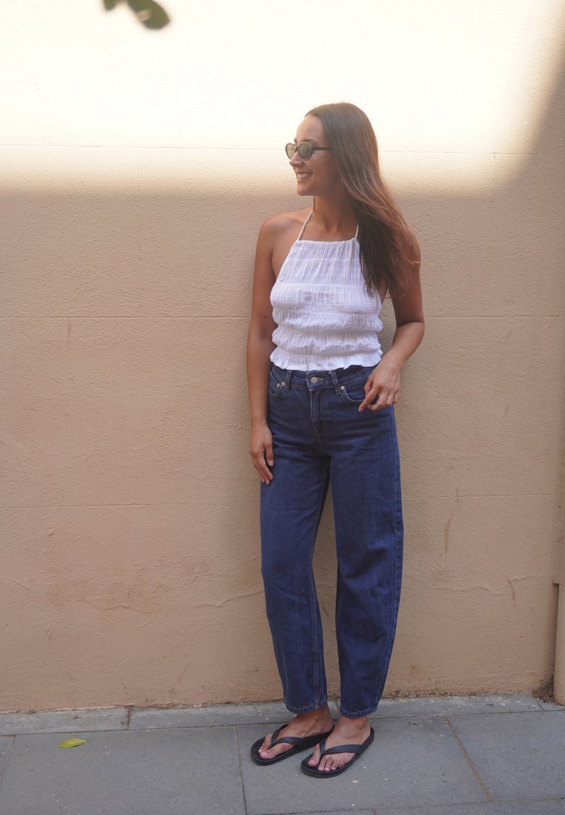 Femme aux longs cheveux bruns portant des lunettes de soleil, un haut blanc sans manches, un jean bleu et des tongs noires, debout contre un mur beige.