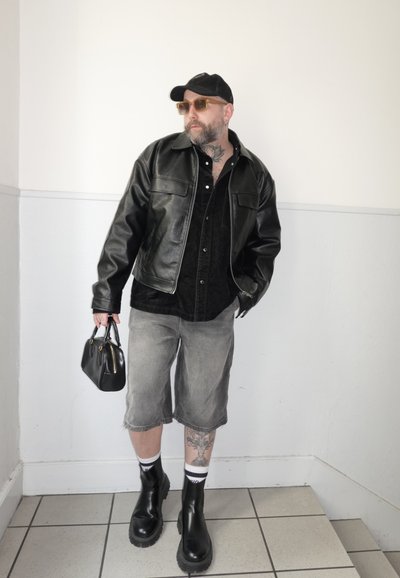Hombre con chaqueta de cuero negra, pantalones cortos grises hasta la rodilla, botas negras, gafas de sol y gorra, sosteniendo un bolso negro contra una pared blanca.