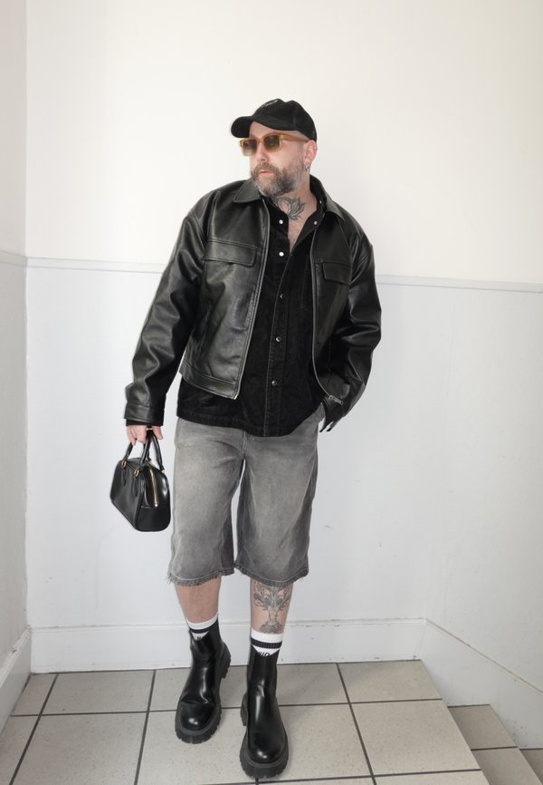 Muškarac u crnoj kožnoj jakni, sivim kratkim hlačama do koljena, crnim čizmama, sunčanim naočalama i kapom, držeći crnu torbu nasuprot bijelog zida.