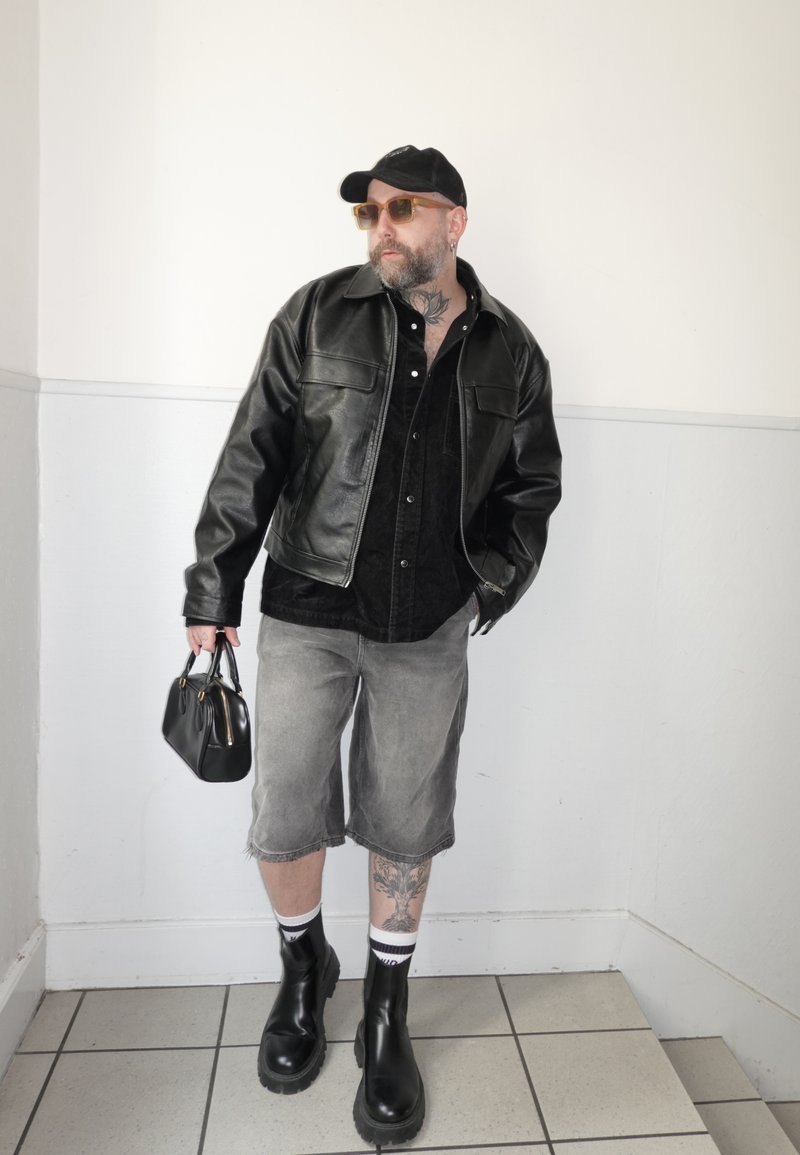 Muž v čiernej koženej bunde, sivých šortkách po kolená, čiernych topánkach, slnečných okuliaroch a čiapke, držiaci čiernu kabelku pri bielej stene.