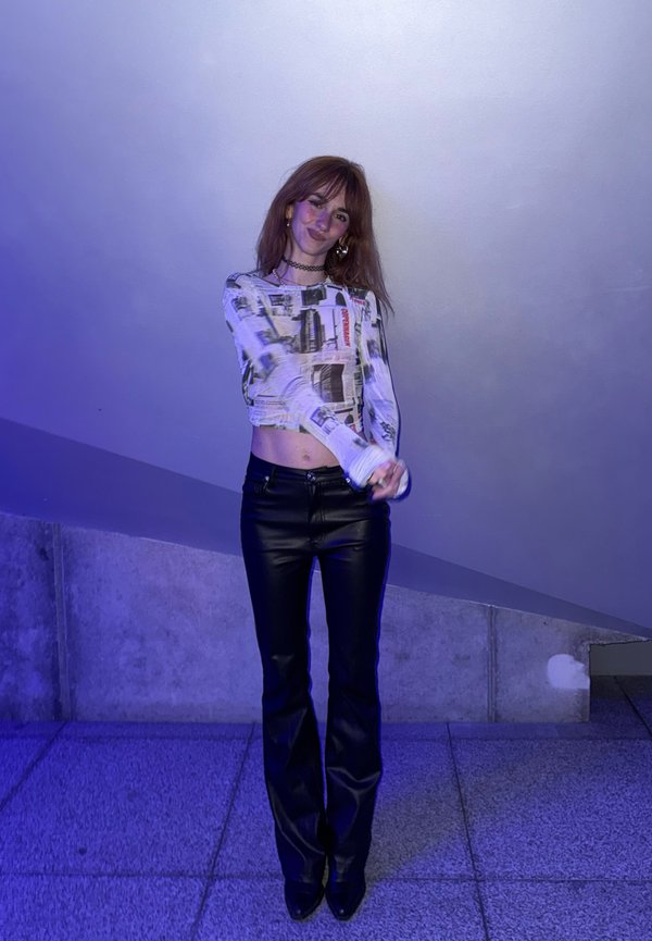 Tânără purtând un crop top cu mâneci lungi cu imprimeu de ziar și pantaloni negri din piele stă sprijinită de un perete înclinat din beton, iluminat cu lumină albastră.