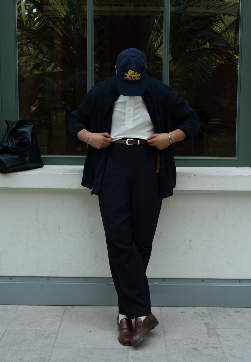Giacca scura su una polo bianca, pantaloni neri con una cintura e scarpe marroni. Un cappellino blu con un logo. Borsa nera accanto. Ambientazione interna.