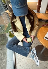 Chaqueta oversized de color amarillo claro, camiseta navy, jeans de denim azul, zapatillas de running plateadas y azules, sosteniendo una bebida verde con diseño de hojas.
