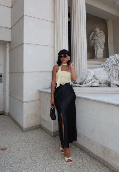 Falda maxi de satén negra con una abertura alta, combinada con un top sin mangas de color amarillo pálido y sandalias con lazo. Un pequeño bolso negro completa el look.