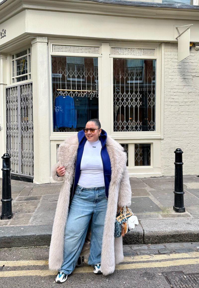 Manteau en fausse fourrure beige sur un sweat à capuche bleu, t-shirt blanc et jean à jambes larges. Tient un petit sac à main. Collines brunes et trottoir visibles.