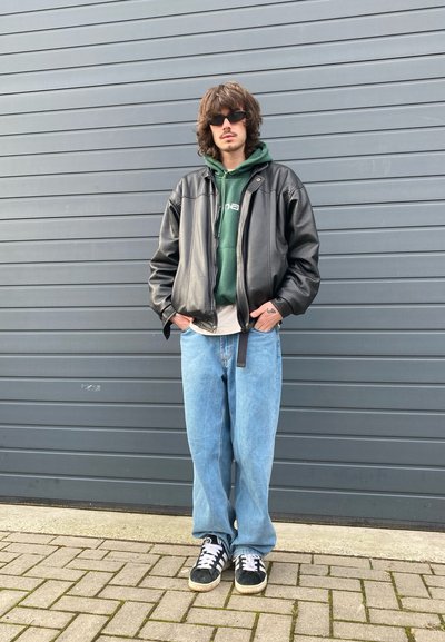 Chaqueta bomber de cuero negra sobre una sudadera con capucha verde, jeans baggy de color azul claro y zapatillas negras con acentos blancos. Unas gafas de sol circulares complementan el look.