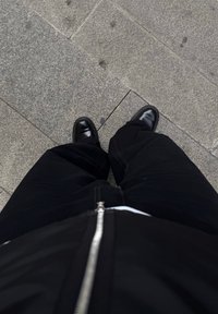 Giacca nera con zip sopra una camicia bianca, abbinata a pantaloni neri e scarpe nere brillanti, vista dall'alto su una superficie di pietra grigia.