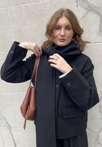 Vrouw met schouderlang rood haar die de kraag van haar donkere jas aanpast, met een bruin leren handtasband in de hand, staand tegen een gestructureerde grijze muur.