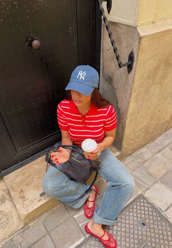Frau mit blauer Kappe, rot gestreiftem Hemd und Jeans, sitzt auf Steinstufen und hält eine Kaffeetasse und eine schwarze Handtasche nahe einer schwarzen Tür.