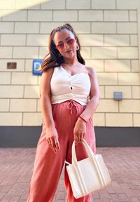 White, buttoned, one-shoulder top; pink, pleated, wide-leg trousers; small white handbag; round pink sunglasses; textured wall background.